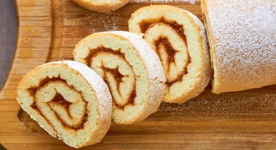 Rotoli di pasta biscotto 3 uova con ripieno di marmellata, spolverati di zucchero a velo su tagliere di legno.