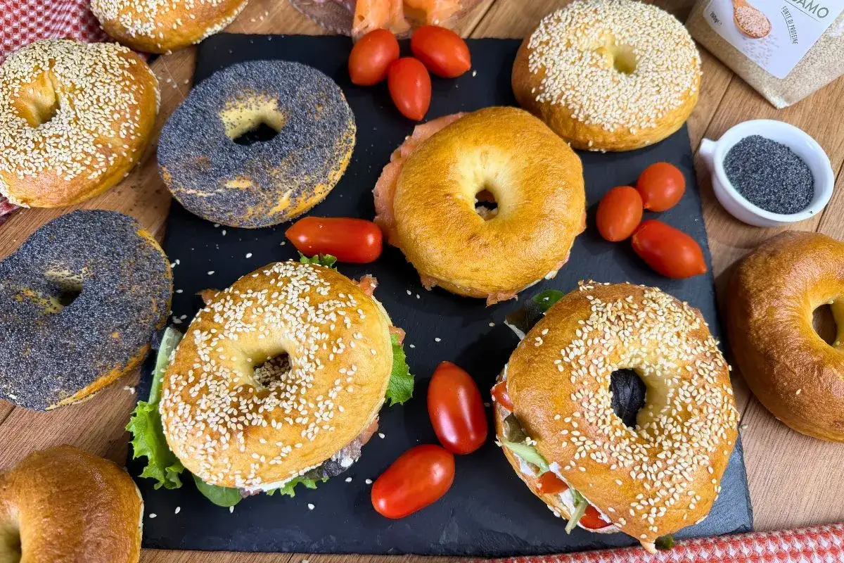 Varietà di bagel dolci e salati, alcuni con semi di sesamo, altri con semi di papavero, accompagnati da pomodorini e salmone.