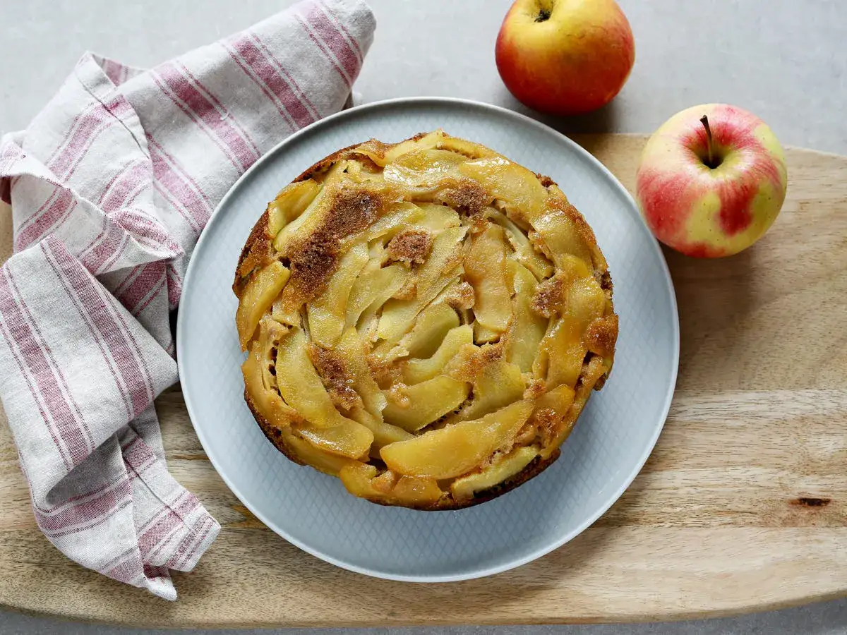 Torta rovesciata di mele e noci, con mele caramellate disposte a spirale e una spolverata di zucchero di canna. Due mele rosse e gialle riposano accanto.
