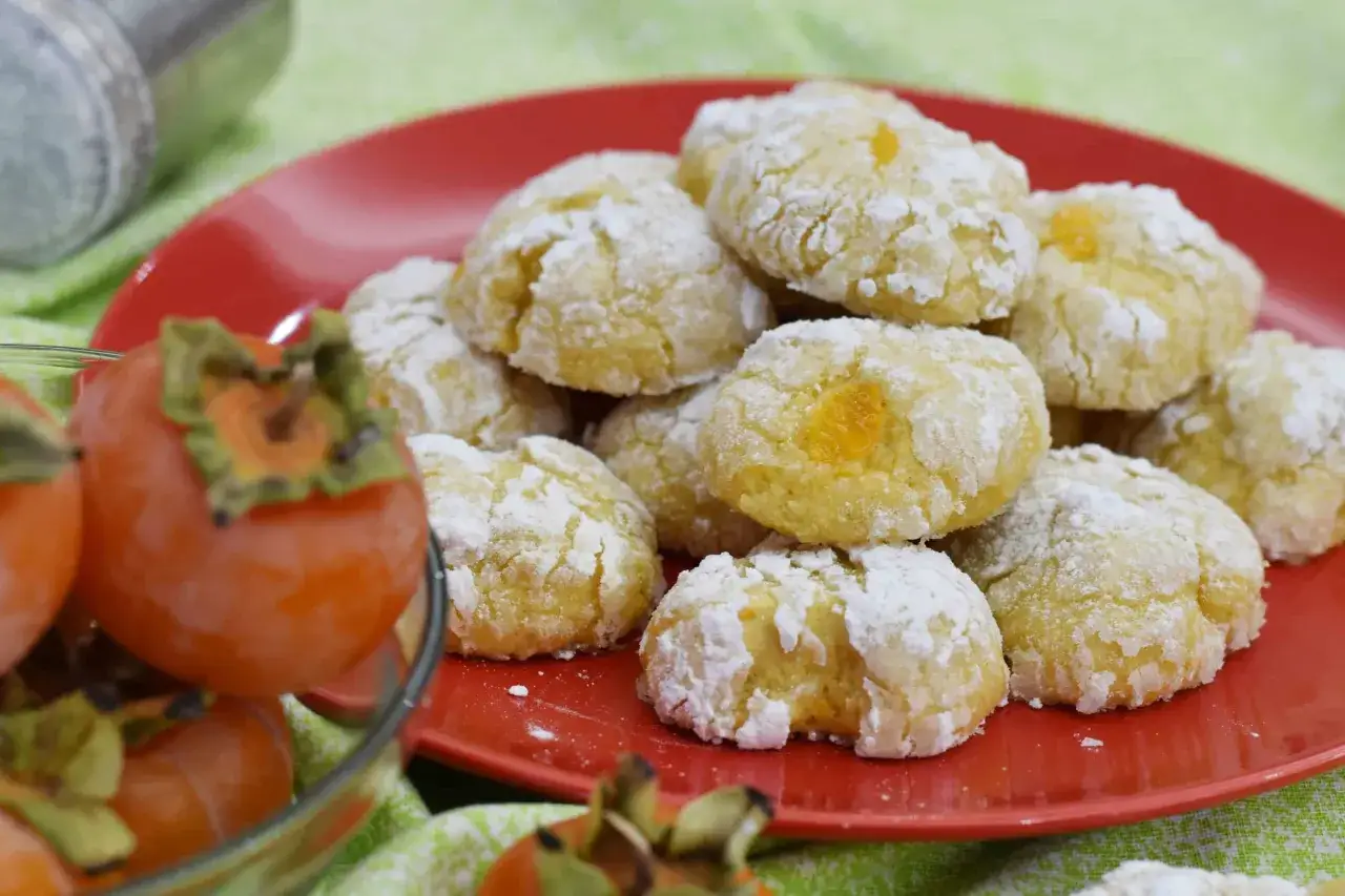 Biscotti ai cachi, dolcetti morbidi e profumati, cosparsi di zucchero a velo, accanto a cachi freschi.