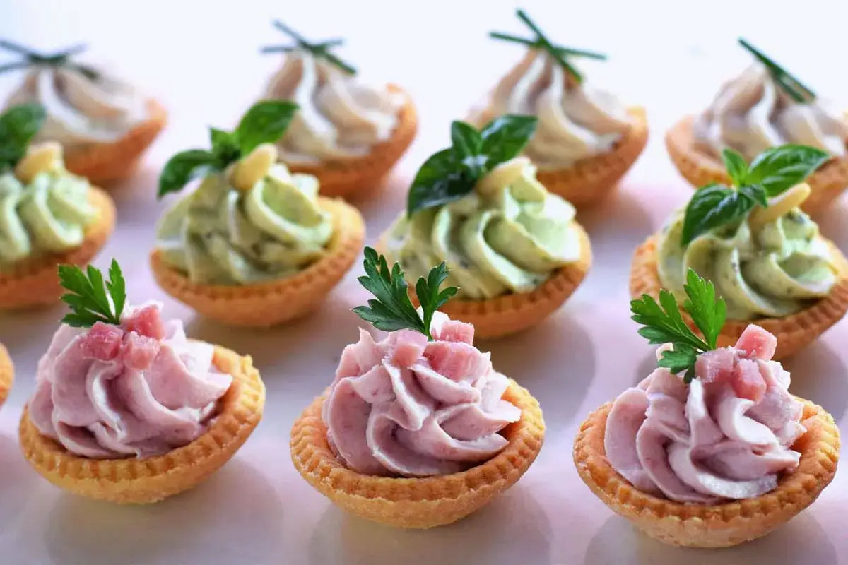 Tartellette con pasta frolla salata con olio e parmigiano, ripiene di crema rosa e verde, guarnite con erbe aromatiche e cubetti di prosciutto.