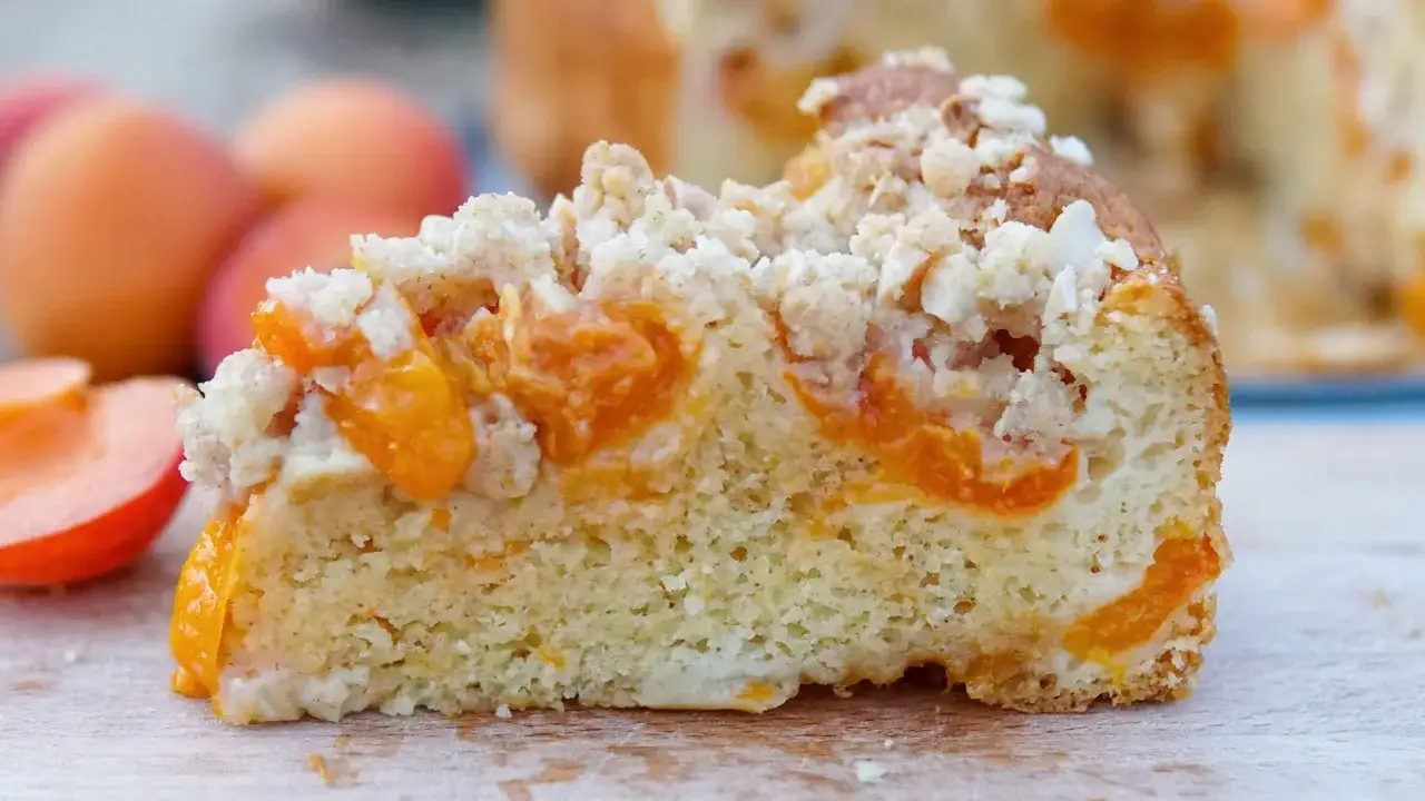 Fetta di ciasto drozdzowe con albicocche e crumble croccante, su uno sfondo sfocato di albicocche fresche.