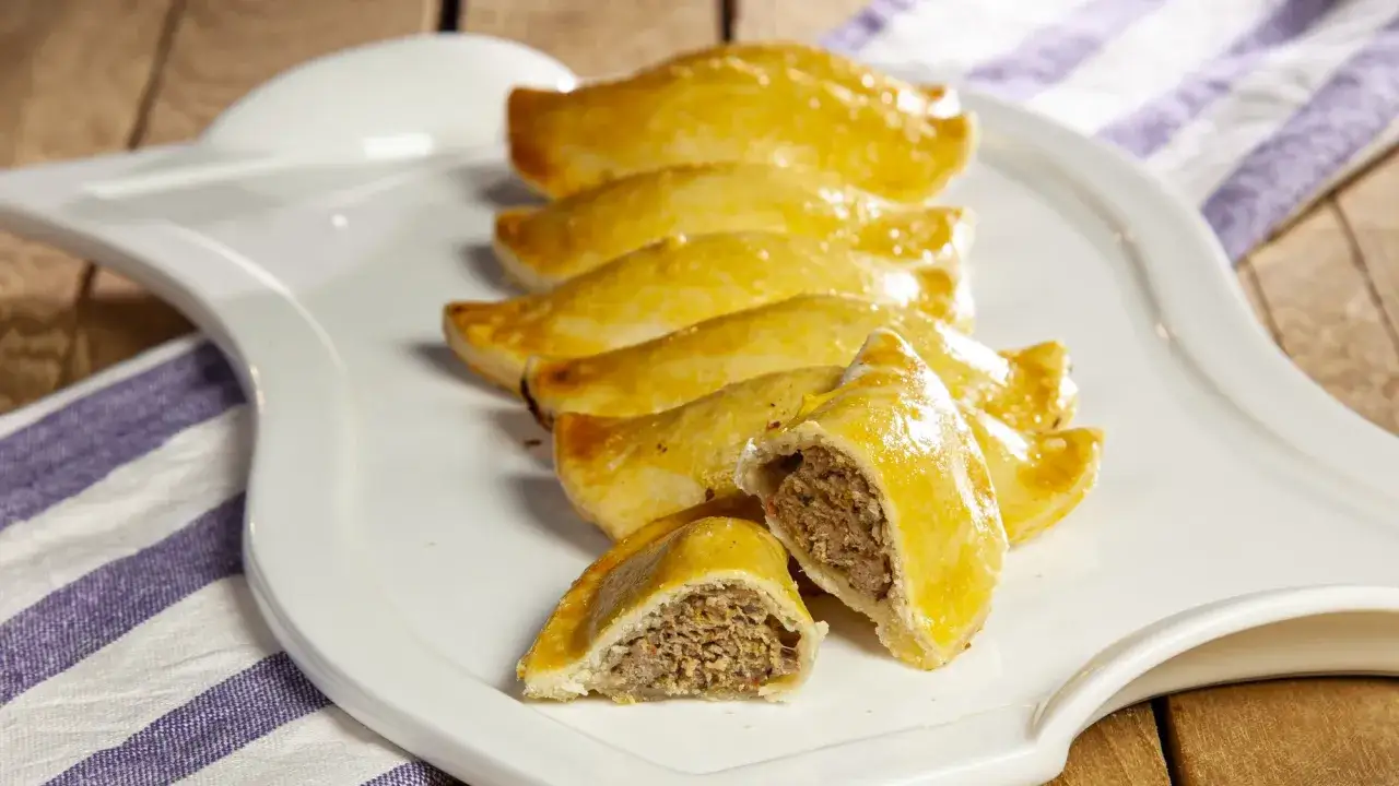 Sette empanadas sarde dorate su un piatto bianco, una aperta rivela un ripieno saporito.