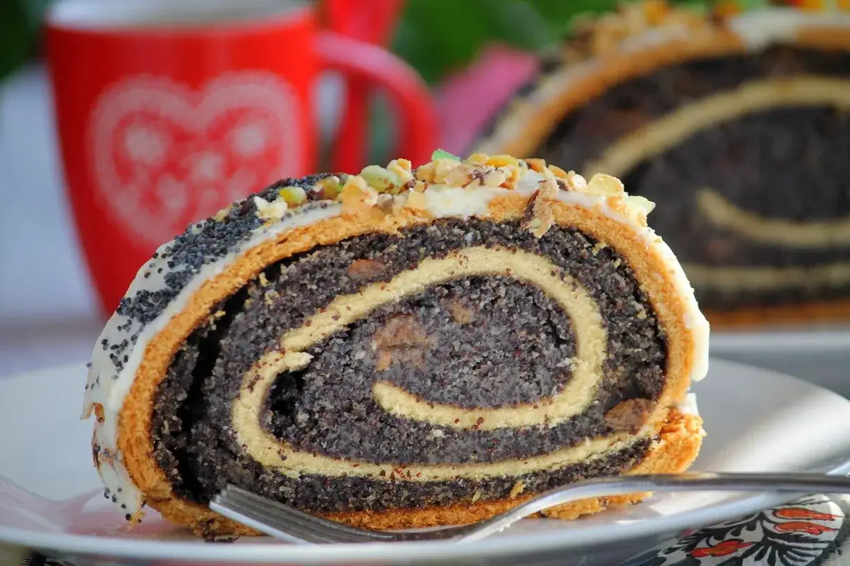 Fetta di makowiec japoński, rotolo di pasta con semi di papavero, glassa bianca e frutta secca, su un piatto bianco con una forchetta.