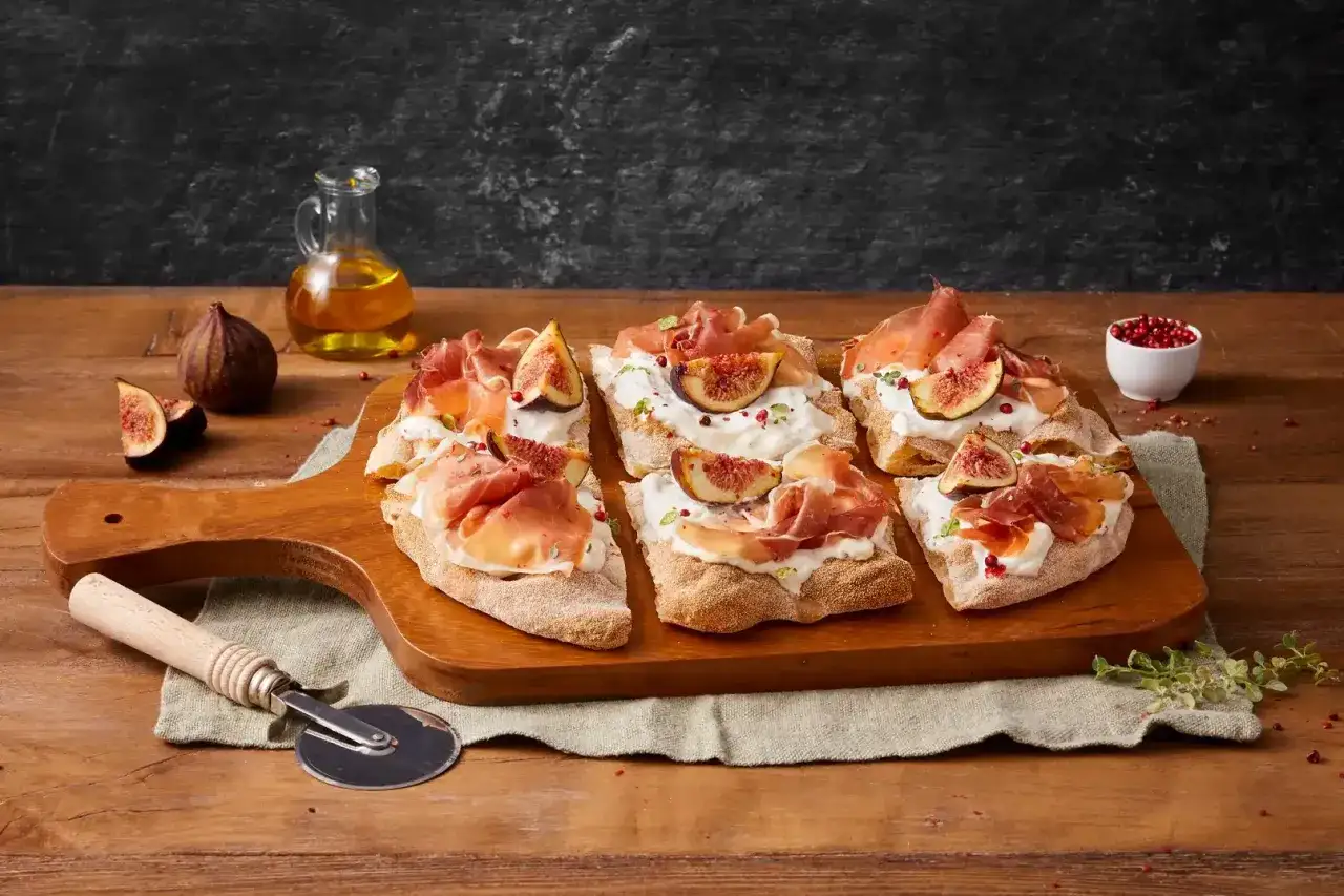 Pinsa crudo e burrata su tagliere di legno, guarnita con fichi freschi e pepe rosa.