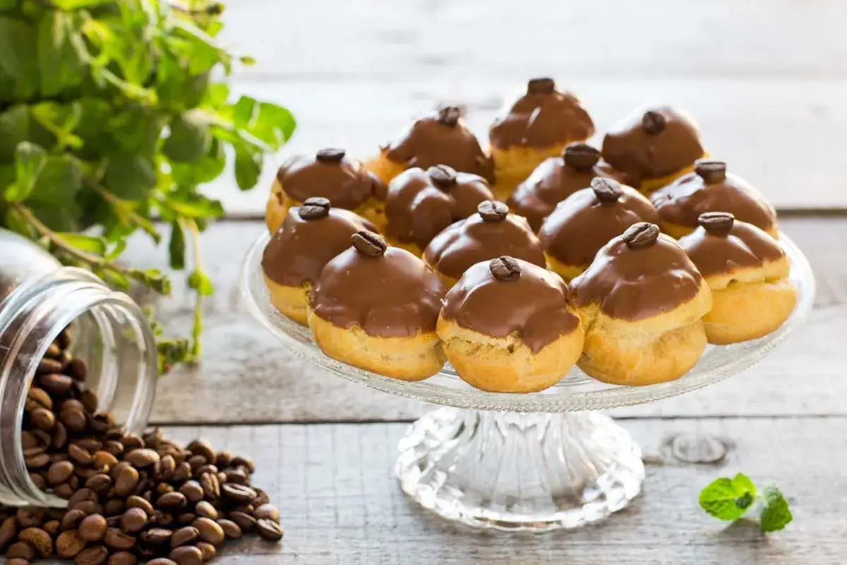 Bign&egrave; al tiramis&ugrave; ricoperti di cioccolato, guarniti con chicchi di caff&egrave;, su un'alzata di vetro, con chicchi di caff&egrave; sparsi e menta fresca.
