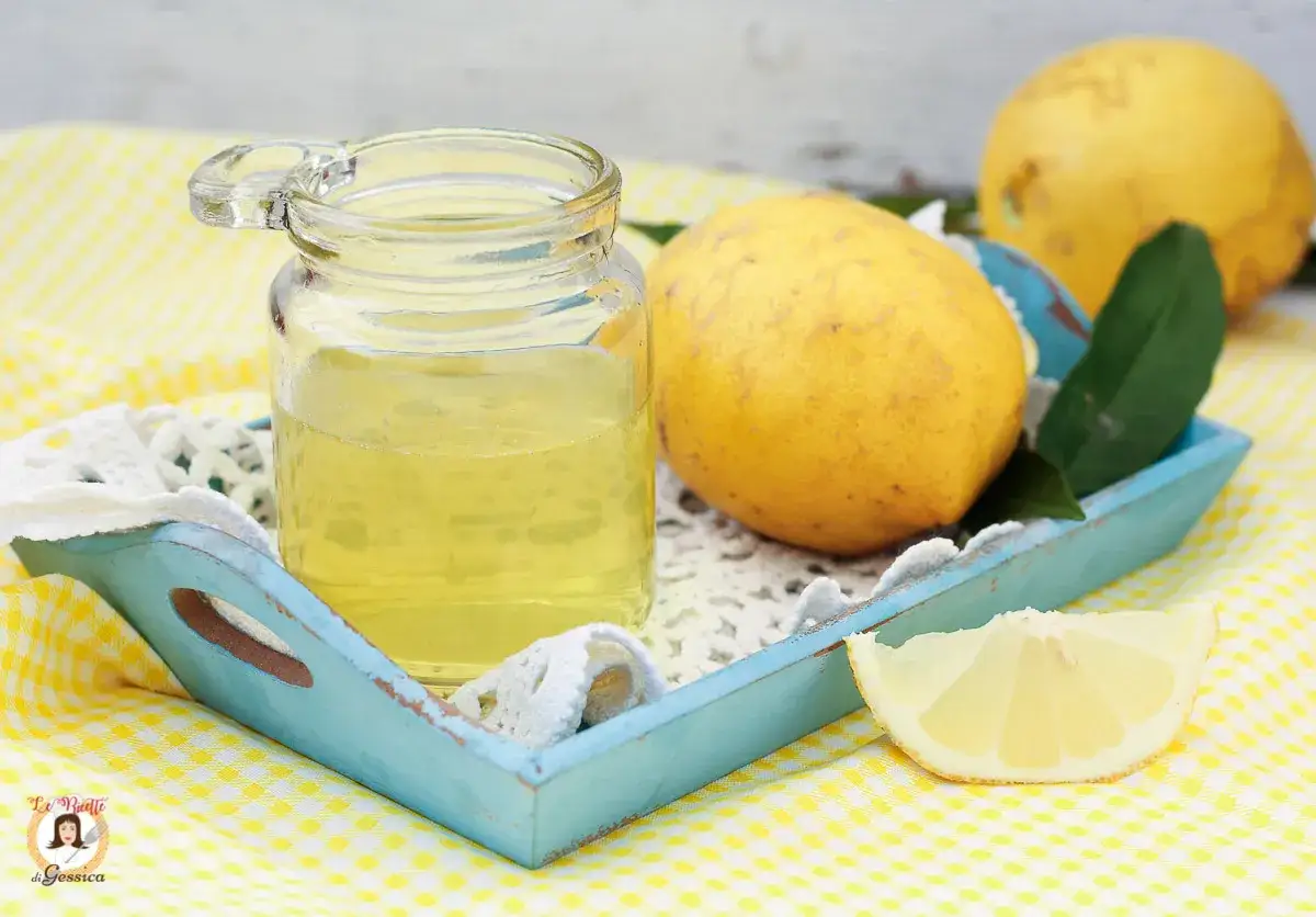 Bagna al limoncello per pan di spagna, con limoni freschi e un vasetto di sciroppo giallo brillante su un vassoio azzurro.