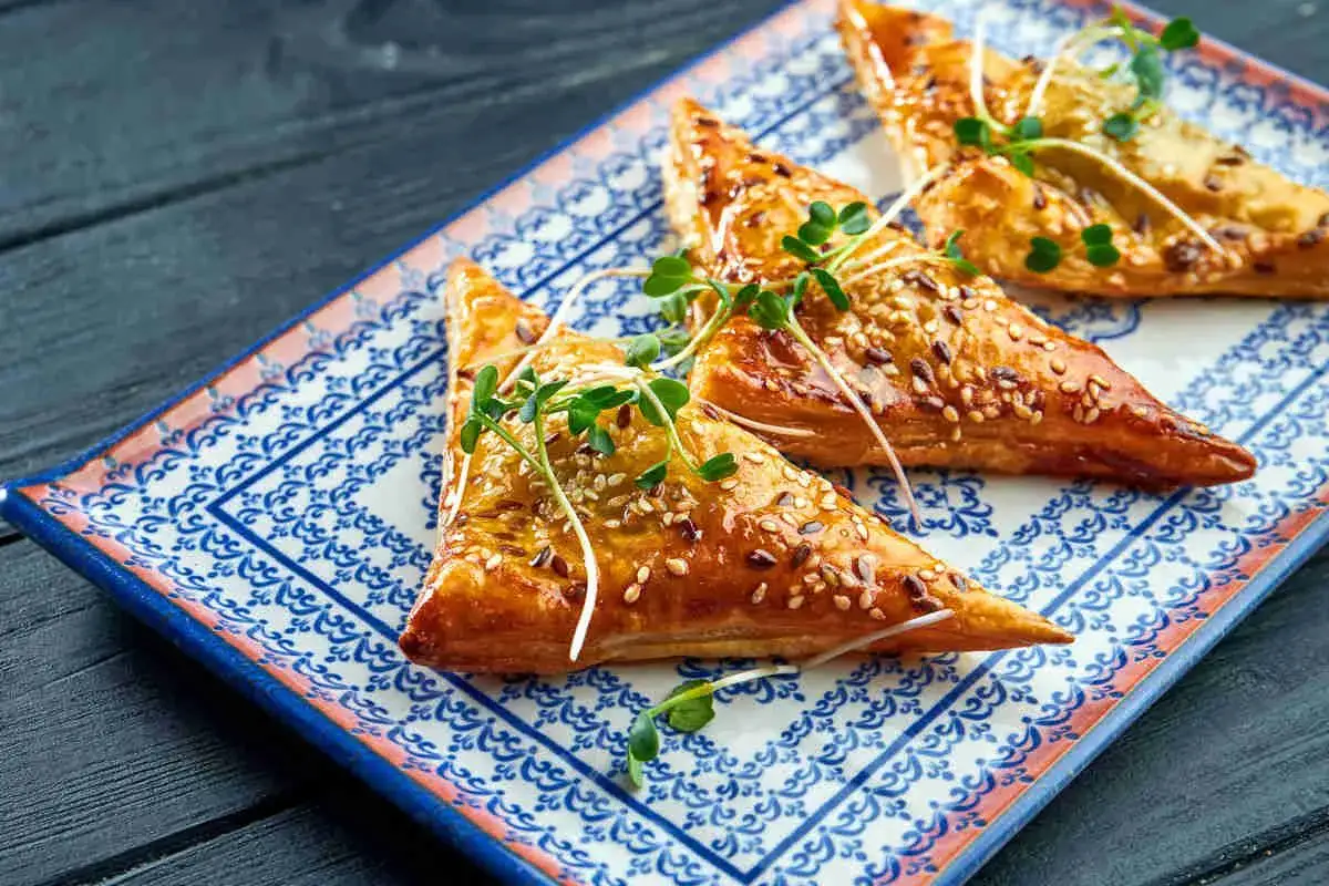 Triangoli dorati di pasta fillo, cotti al forno e cosparsi di semi di sesamo, guarniti con germogli freschi su un piatto decorato.