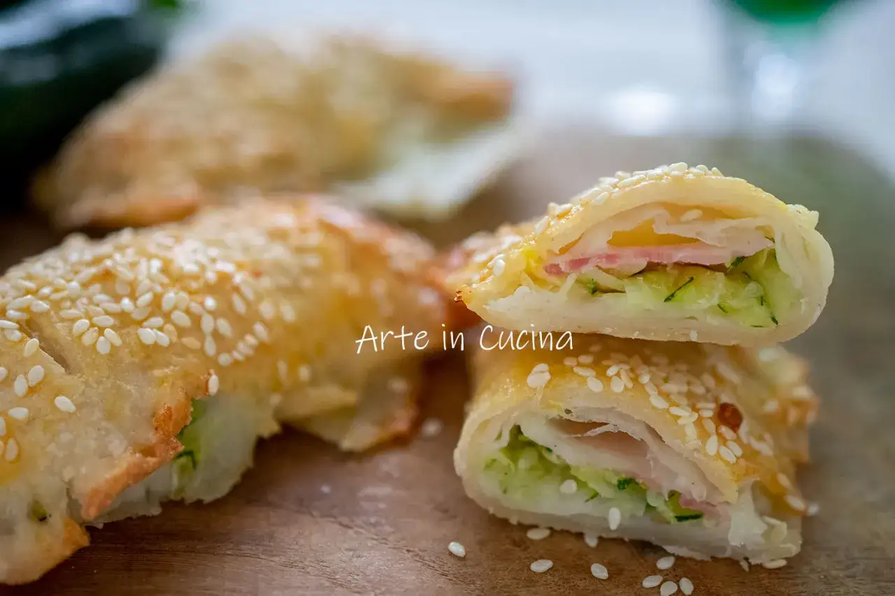 Fagottini di pasta sfoglia dorata con zucchine, scamorza filante e prosciutto, cosparsi di semi di sesamo.