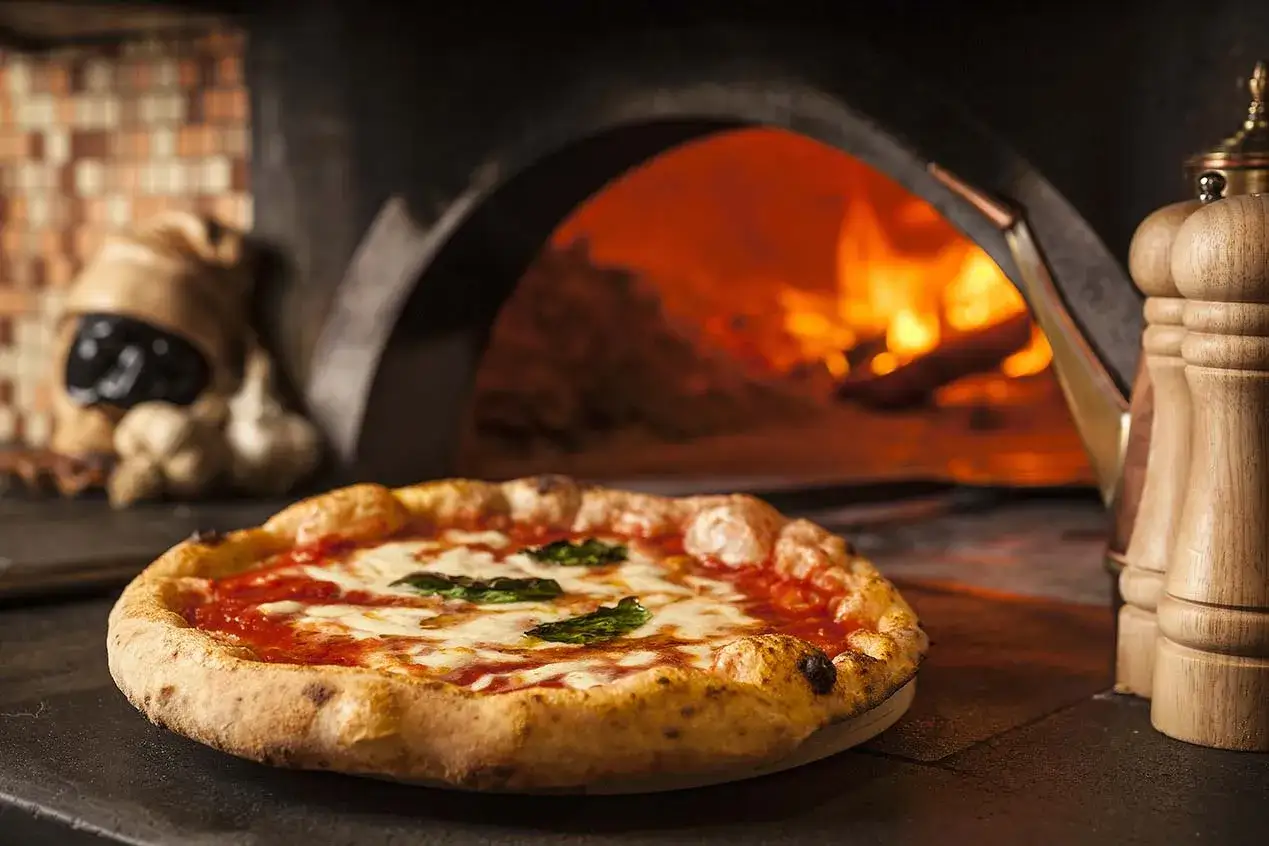 Pizza napoletana appena sfornata, con pomodoro, mozzarella filante e basilico, accanto a un forno a legna ardente.