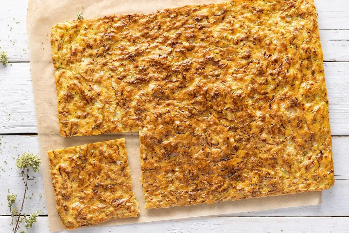 Schiacciata di zucchine e cipolle dorata e croccante, tagliata a pezzi su carta da forno.