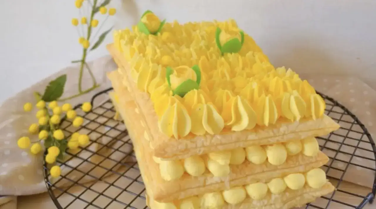 Tronchetto mimosa, un dolce a strati di pasta sfoglia e crema gialla, decorato con ciuffi di crema e fiorellini verdi, che ricorda la primavera.