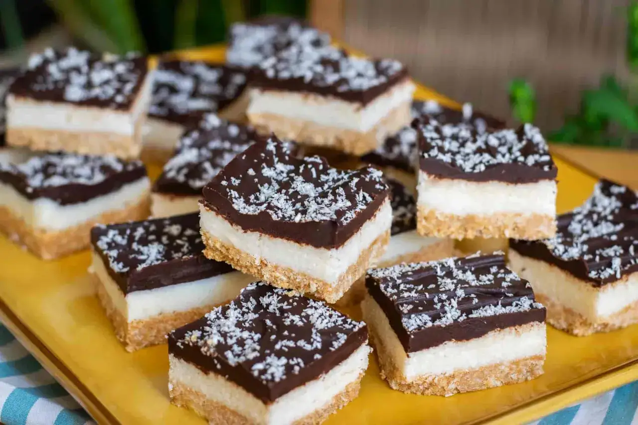 Deliziosi quadrotti cioccolato e cocco, stratificati con base croccante, crema bianca e copertura fondente, spolverati con cocco grattugiato.