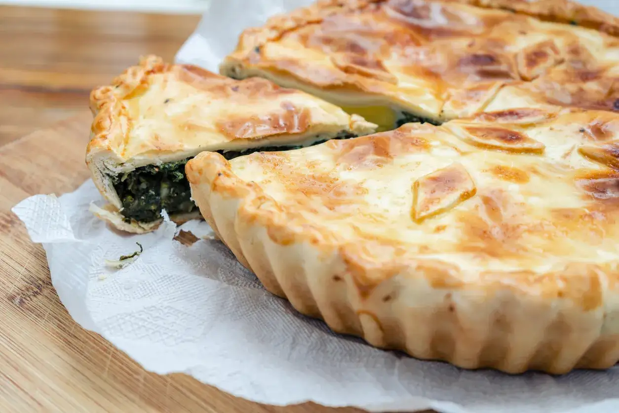 Torta pasqualina senza uova, con ripieno verde e crosta dorata, tagliata a fette su carta bianca.