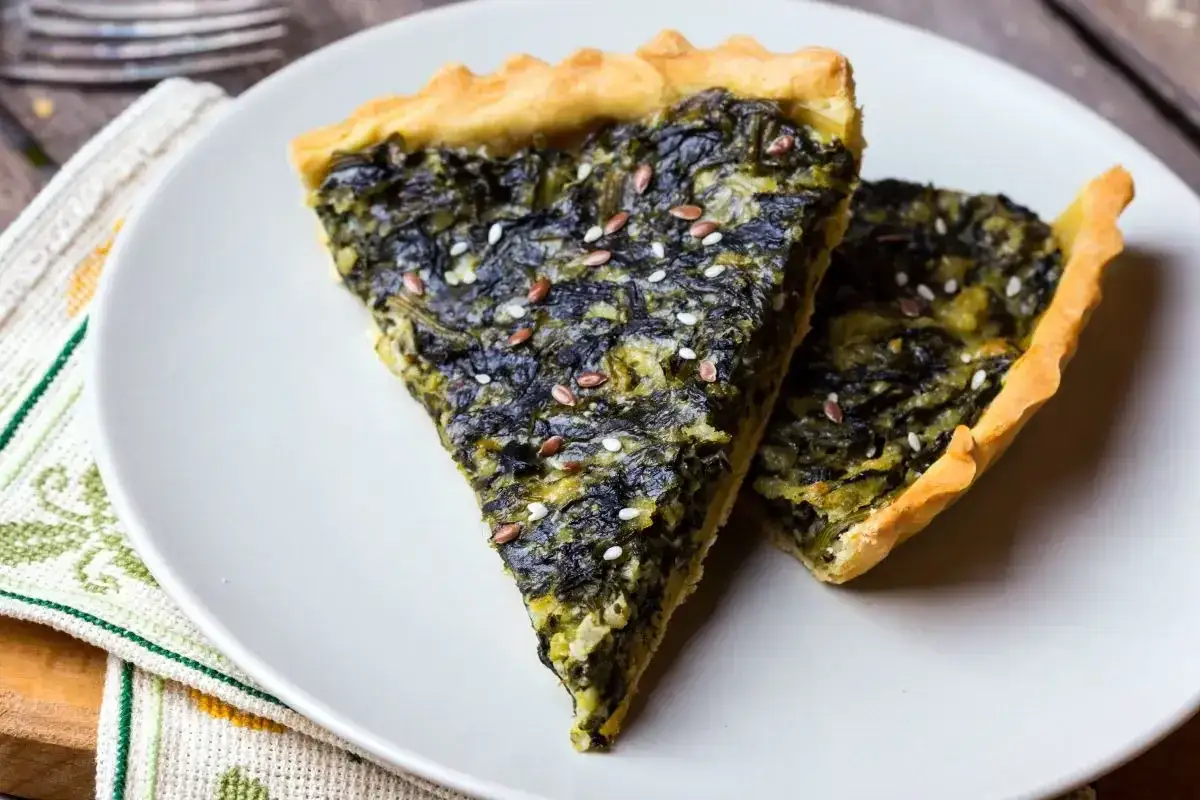 Fette di torta salata vegana spinaci, guarnite con semi di lino e sesamo, su un piatto bianco.