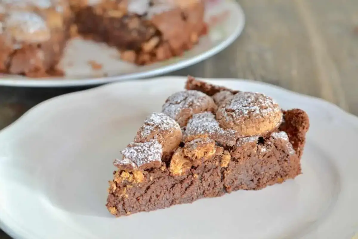 Una fetta di torta amaretti e cioccolato fondente, cosparsa di zucchero a velo, su un piatto bianco.