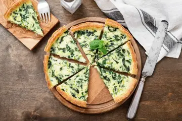 Torta salata spinaci e mozzarella, con un tocco di speck affumicato, tagliata a spicchi su un tagliere di legno.