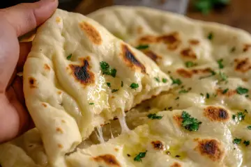 Una mano strappa un pezzo di naan al formaggio filante, cosparso di prezzemolo fresco e burro fuso.