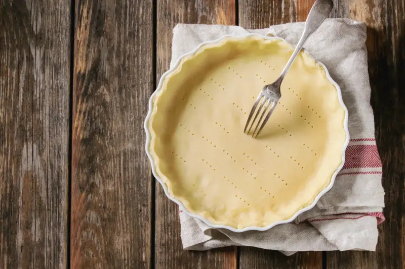 Pasta frolla salata con olio e parmigiano pronta per essere infornata. Una forchetta è appoggiata sulla base bucherellata.